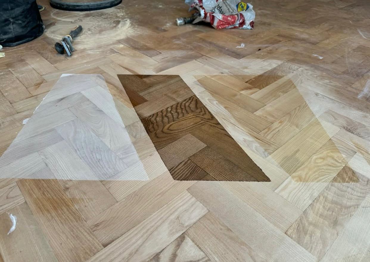Wood Joyful Restoration in Lee: stain-testing swatches on a sanded oak herringbone parquet (solid wood) floor showing light, medium and dark finish options.